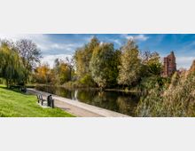 Parklandschaft mit einem kleinen Fluss, umgeben von Bäumen in herbstlichen Farben. Im Vordergrund eine Holzpromenade mit Sitzbänken, im Hintergrund ein altes Backsteingebäude.