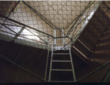 Blick nach oben in einem Treppenhaus mit Metallgeländern und Zwischengittern, umgeben von einem engmaschigen Sicherheitsnetz. Licht fällt durch ein Dachfenster und beleuchtet die Metallkonstruktion.