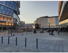 Moderner urbaner Platz mit gepflastertem Boden, umgeben von Glas- und Metallgebäuden. Im Hintergrund ist ein großes Gebäude mit spiegelnder Fassade und einem Mercedes-Logo zu sehen, während Bauarbeiten auf einer abgesperrten Fläche stattfinden.