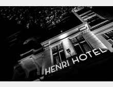 Nachtsicht eines historischen Hotelgebäudes mit Schriftzug „Henri Hotel“ in leuchtenden Buchstaben, großen Fenstern und verzierter Fassade; darüber sind Baumzweige im Dunkeln sichtbar.