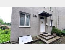 Eingangsbereich eines Wohngebäudes mit grauer Putzfassade, einer weißen Haustür mit Dekoration, und einem kleinen Vordach. Links und rechts der Tür befinden sich Fenster, und vor dem Eingang führt eine kurze Steintreppe mit Blumendekoration im Topf zum Eingang.