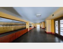 Langer Flur mit gelb gestrichenen Wänden und einer Garderobe auf der linken Seite mit zahlreichen Haken. Rechts sind Türen mit Glaselementen und ein Heizkörper sichtbar, der Boden ist dunkel gefliest.