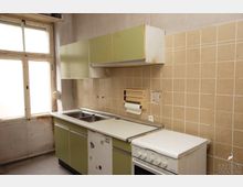 Eine kleine, veraltete Küche mit grünlichen Oberschränken, beigen Fliesen an der Wand und einem weißen Elektroherd. Links befindet sich ein großes Fenster mit sichtbaren Gebrauchsspuren am Rahmen.