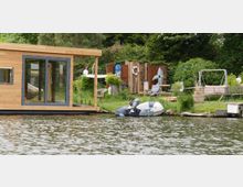 Ein schwimmendes Holzhaus mit großen Glasfenstern steht am Ufer eines Sees. Im Hintergrund sind ein Gartenbereich mit einer Sitzbank, einem Schuppen und einem Schlauchboot erkennbar, umgeben von dichter Vegetation.