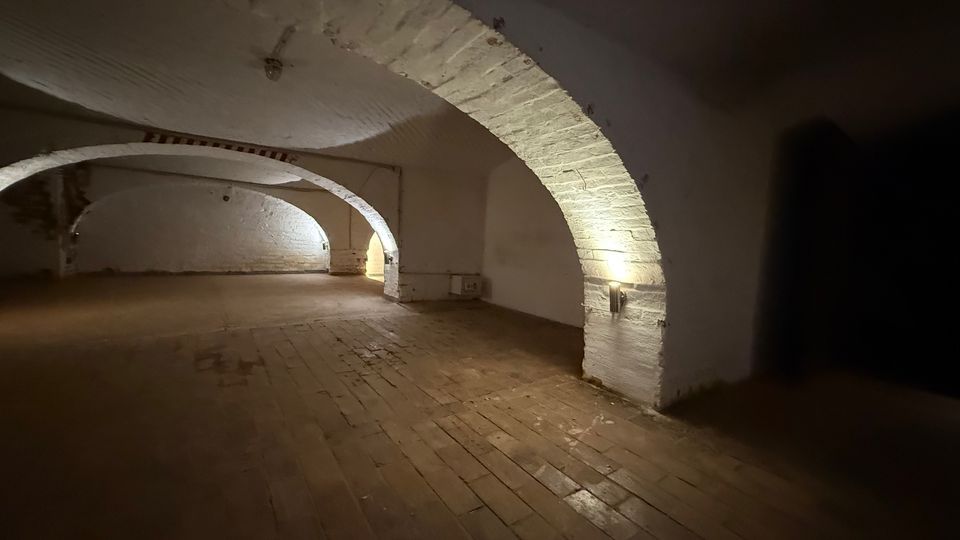 Großer, leerer Keller- oder Gewölberaum mit weiß verputzten Backsteinbögen und niedrigem Tonnengewölbe. Holzplankenboden und wenige Wandlampen beleuchten die gebogenen Durchgänge und Nischen.