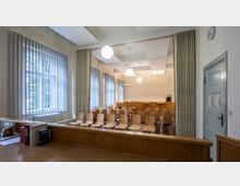 Ein Konferenz- oder Gerichtssaal mit Holzstühlen in Reihen, einer erhöhten Richterbank und großen Fenstern mit vertikalen Jalousien. An der Wand hängen Kugellichter, und eine graue Tür befindet sich rechts im Bild.