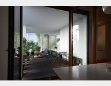 Überdachte Terrasse mit Holzboden, ausgestattet mit einem Tisch, Stühlen und mehreren Topfpflanzen; im Hintergrund ist eine Treppe und Begrünung entlang der weißen Wände zu sehen.