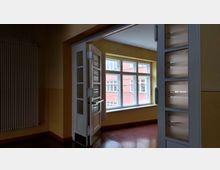 Blick in einen sonnigen Raum mit gelb gestrichenen Wänden, großen weißen Fenstern und einer teilweise offenen weißen Tür mit Milchglaselementen. Durch die Fenster ist die Fassade eines angrenzenden Gebäudes in Rosa sichtbar.