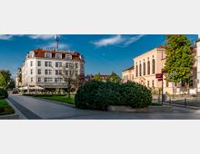 Städtischer Platz mit einem mehrstöckigen Gebäude links, das „Hotel Kaiserhof“ mit rotem Ziegeldach und umliegendem Straßencafé zeigt, sowie einem klassizistischen Gebäude mit großen Rundbogenfenstern rechts. Im Vordergrund befinden sich Wege, Grünflächen und Büsche.