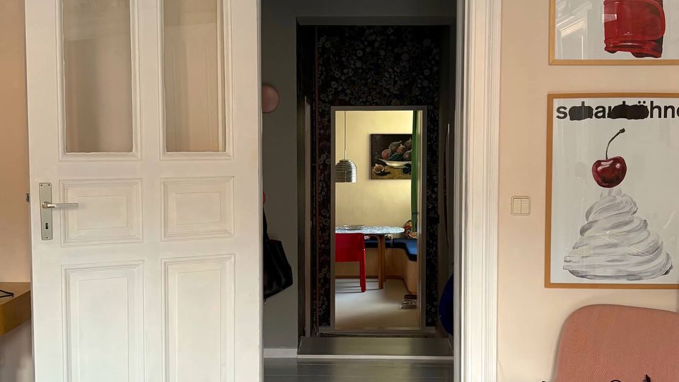 Interior of a home with pale walls, white-painted wood floors, and an ornate white doorway with decorative molding and a partially open double door leading to a darker hallway and another room beyond. On the right wall are two framed posters, and a low chair sits near the corner.