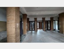 Heller Eingangshallenbereich mit Säulen aus poliertem Stein, einer Wand mit Holztüren mit Glaseinsätzen und schmuckvoll gestaltetem Deckengesims. Der Boden besteht aus großen Steinfliesen, und im Hintergrund ist eine geöffnete Doppeltür zu sehen.