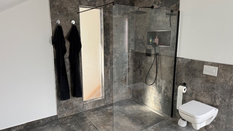 Modern bathroom with large gray stone-look tiles, a walk-in glass shower with rainfall showerhead and recessed shelf, and a wall-mounted toilet. A tall mirror and towel hooks are on the left wall, with white upper walls and small ventilation grilles near the ceiling.