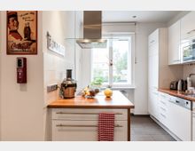 Moderne helle Küche mit weißen Schränken und Holzarbeitsplatten. Ein zentrales Fenster bietet Blick nach draußen, während ein Küchenblock mit rotem Geschirrtuch, Obst und einer Saftpresse im Vordergrund steht.