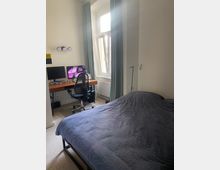 Schlafzimmer mit einem Schreibtisch und einem Computer auf der linken Seite, einem schwarzen Bürostuhl davor und einem Bett mit dunkelblauer Bettdecke im Vordergrund. Helles Tageslicht fällt durch ein großes Fenster mit langen, hellgrünen Vorhängen.