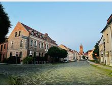 Kopfsteingepflasterte Straße in einer Kleinstadt mit historischen Backstein- und Putzhäusern, darunter ein Eckgebäude mit Hotelbeschilderung. Im Hintergrund ist ein roter Kirchturm mit Spitze am Ende der Straße zu sehen.