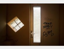 Gelb gestrichene Innenseite einer Tür mit einem langen schmalen Fenster und Graffiti-Schriftzügen. Links daneben befindet sich ein schräg eingebautes quadratisches Fenster mit milchigem Glas.