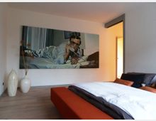 Schlafzimmer mit Doppelbett und einer orangefarbenen Bank am Fußende, hellem Holzboden und einem großen Gemälde eines Mannes am Telefon an der Wand. Daneben stehen zwei dekorative weiße Vasen mit getrockneten Pflanzen.