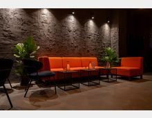 Moderne Lounge mit orangefarbenen Sofas, schwarzen Sesseln und niedrigen, schwarzen Metalltischen, dekoriert mit Glasflaschen und Gläsern. Im Hintergrund eine strukturierte, dunkelgraue Wand, beleuchtet von mehreren Spotlights, neben grünen Zimmerpflanzen.