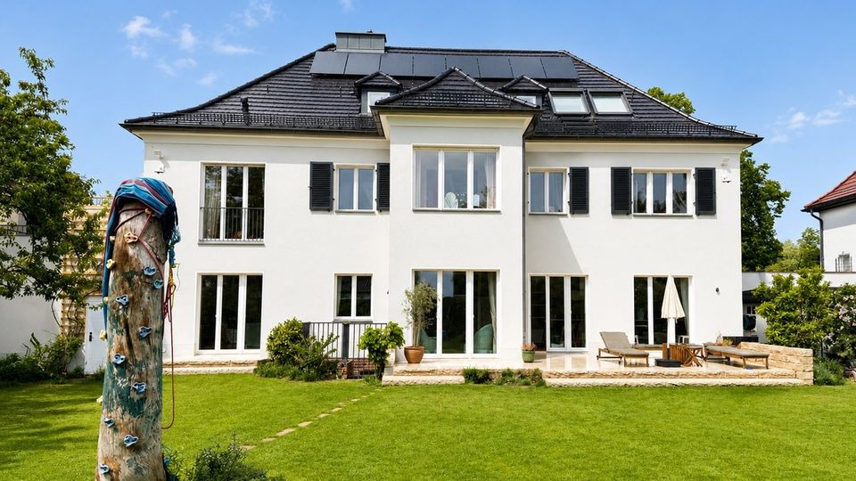 Großes weißes Wohnhaus mit dunklem Walmdach, Dachfenstern und Solarpaneelen, symmetrischer Fensterfront mit schwarzen Fensterläden und mehreren Terrassentüren. Davor liegt ein weitläufiger Rasen mit Terrasse und Gartenmöbeln sowie einem Kletterstamm am linken Bildrand.