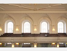 Innenansicht einer historischen Kirche mit weißen Wänden, großen, hochgewölbten Fenstern und dekorativen Stuckverzierungen. Im oberen Bereich befinden sich hölzerne Sitzbänke auf einer Empore, die von Wandlampen beleuchtet wird.