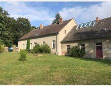 Ein ländliches Haus mit grauem Putz und rotem Ziegeldach, ergänzt durch mehrere Sprossenfenster und eine Dachgaube mit Glasfläche. Das Gebäude befindet sich in einem grasbewachsenen Garten mit Bäumen im Hintergrund und einem geparkten Fahrzeug links neben einer Holztoröffnung.