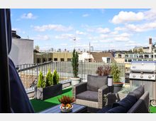Dachterrasse mit Sitzmöbeln aus Geflecht, einem großen Gasgrill und dekorativen Pflanzen in Töpfen und Pflanzkästen, mit Blick auf umliegende Gebäude und den Berliner Fernsehturm am Horizont unter blauem Himmel.