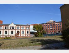 Historische Backsteingebäude mit weißen und roten Fassadenelementen, teils modernisiert, in einem offenen Hofbereich mit kleiner Grünfläche und einzelnen Bäumen. Der Himmel ist klar und blau, und im Hintergrund sind parkende Autos zu sehen.