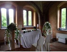 Eine historische Innenraumkulisse mit hohen Fenstern, einem gewölbten, bemalten Deckenbogen und verputzten Wänden. Im Zentrum steht ein dekorierter Tisch mit weißer Tischdecke, Kerzenständern und Blumengirlanden, davor zwei weiß überzogene Stühle.