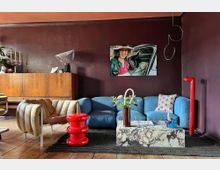 Wohnzimmer mit violett gestrichener Wand, einem blauen Sofa, einem beigefarbenen Lounge-Sessel, einem roten Beistelltisch und einem rechteckigen Couchtisch aus Marmor. Ein Holzschrank, ein Foto an der Wand und dekorative Accessoires wie Blumenvasen und eine Tischlampe prägen den Raum.