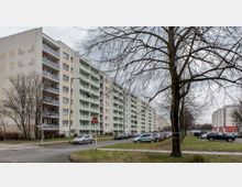 Mehrstöckige Wohngebäude in Plattenbauweise mit hellen Fassaden und Balkonen, links ein kahler Baum und rechts eine parkende Autoschlange entlang der Straße. Der Himmel ist bewölkt, und die Szene wirkt winterlich.