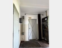 Ein schlichter Technikraum mit einer Metalltür in der Mitte, über der ein kleines Schild mit der Aufschrift "Hbw Blok 1189" hängt. Links befinden sich Rohre und eine Elektroeinheit an der Wand, rechts ein Schaltschrank mit sichtbaren Schaltern und Verkabelungen.