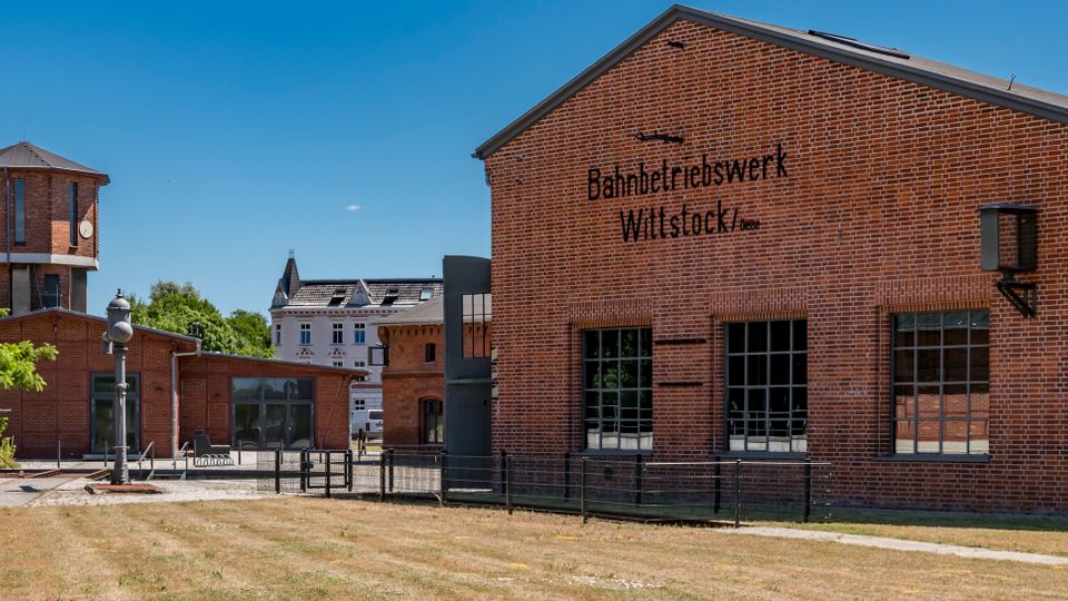 Backsteingebäude eines Bahnbetriebswerks mit großen Sprossenfenstern und der Aufschrift „Bahnbetriebswerk Wittstock“, davor eine Grasfläche und ein umzäunter Hof. Links steht ein markanter, runder Backstein-Wasserturm mit Zeltdach; im Hintergrund sind weitere Gebäude und Grünflächen zu sehen.
