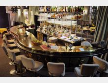Eine stilvolle Bar mit geschwungener glänzender Theke, umgeben von gepolsterten Drehstühlen. Im Hintergrund sind Spiegel, Regale mit Flaschen und Gläsern, sowie Vorhänge sichtbar.