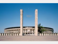 Breite Ansicht des Berliner Olympiastadions mit zwei hohen Steinpylonen, zwischen denen die olympischen Ringe hängen. Das Stadion hat eine halbkreisförmige Säulenfassade, und der Vorplatz ist mit Steinplatten gepflastert.