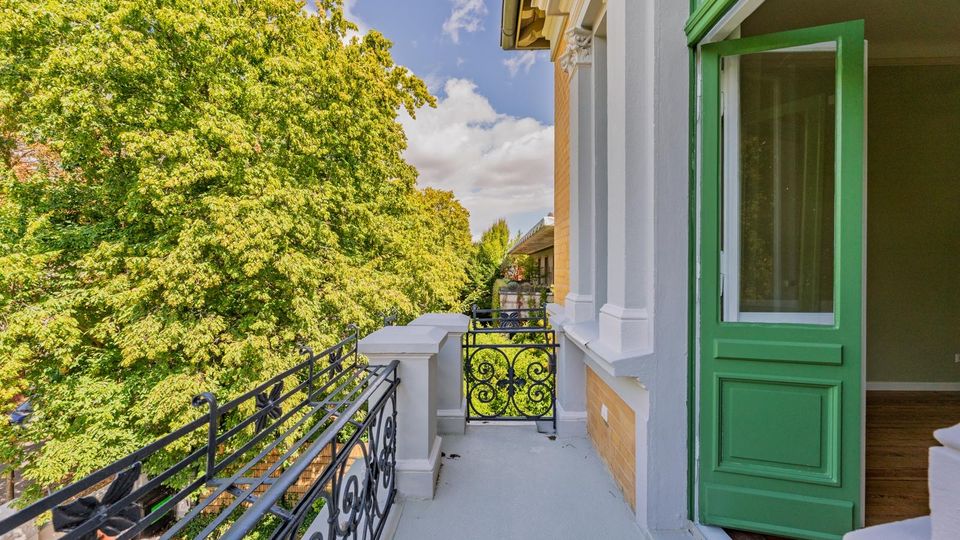 Schmaler Balkon an der Fassade eines Wohnhauses mit weißer Stuckverkleidung, Ziegelwand und schwarzem schmiedeeisernem Geländer. Eine offene grüne Balkontür führt in den Innenraum; neben dem Balkon stehen hohe, dicht belaubte Bäume.
