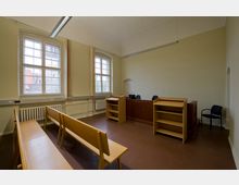 Ein kleiner, einfacher Gerichtssaal mit Holzbänken für Zuschauer, einem erhöhten Richtertisch aus Holz, zwei Rednerpulten und großen Fenstern mit Sprosseneinteilung. Die Wände sind cremefarben, der Boden braun, und Licht fällt durch die Fenster ein.