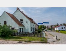 Straßenszene in einer Wohngegend mit freistehenden Einfamilienhäusern, die ein Satteldach haben, und Vorgärten. Im Vordergrund steht ein Straßenschild mit den Namen „Am Weinberg“ und „Schwanebecker Weg“, daneben ein kleines, mit Gras bewachsenes Grundstück.