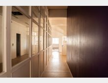 Langer, schmaler Flur in einem leeren Bürogebäude mit Glaswänden und weißen Rahmen auf der linken Seite und einer dunklen Wand rechts. Am Ende des Ganges fällt Tageslicht durch Fenster in einen offenen Raum; der Boden wirkt abgenutzt und die Decke ist teilweise geöffnet.