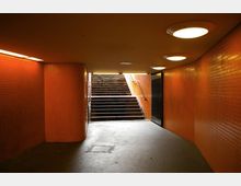 Unterführung mit orangefarbenen Fliesen an den Wänden, beleuchtet durch runde Deckenlampen, führt zu einer Treppe, die nach draußen ins Tageslicht führt. Die Umgebung wirkt schlicht und funktional.