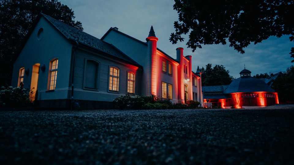Niedrige Außenansicht eines großen, hellen Gebäudekomplexes mit steilem Dach und mehreren säulenartigen Türmen, deren Fassaden bei Dämmerung rot angestrahlt sind. Daneben steht ein rundes Nebengebäude mit Kuppeldach, ebenfalls rot beleuchtet, umgeben von Bäumen und einem dunklen Hofbereich im Vordergrund.