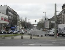 Straßenszene in einer Stadt mit mehreren Fahrstreifen, Ampeln und parkenden Autos. Links befinden sich Gebäude mit roten Fahnen, rechts Werbetafeln und Hinweisschilder, darunter eines mit der Aufschrift „Messegelände ICC“. Im Hintergrund sind Bäume und weitere Gebäude sichtbar.
