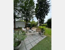 Gartenbereich mit einer Terrasse aus grauen Platten, möbliert mit Sitzgruppe aus hellen Sofas und weißen Stühlen. Umgeben von hohen Bäumen, Rasenflächen und dekorativen Pflanzen in Töpfen, darunter eine Palme und ein geschlossener Sonnenschirm.