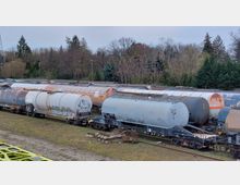 Mehrere Kesselwaggons stehen auf einem Güterzugabstellgleis, teils mit Graffiti versehen. Im Hintergrund sind Bäume und einige Büsche zu sehen, die eine grüne Umgebung andeuten.