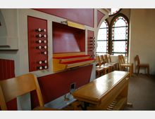 Innenraum einer Kapelle mit einer Kirchenorgel im Vordergrund. Die Orgel verfügt über rote und cremefarbene Verkleidung, mehrere Registerzüge und eine hölzerne Pedaleinheit; im Hintergrund sind Stühle sowie bunte Bleiglasfenster zu sehen.