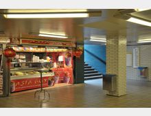 Im Bild ist ein kleiner asiatischer Imbissstand namens "Asia Box" in einer hell beleuchteten U-Bahn-Passage zu sehen. Der Stand ist mit roten Dekorationen geschmückt, darunter Laternen, und bietet Speisen in einer Vitrine an; im Hintergrund sind bläulich geflieste Wände und eine Treppe sichtbar.