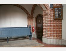 Kirchenecke mit roten Ziegelbogen und einem Holzpodest. Ein blau gestrichener Holzbänksitz steht links, daneben eine hölzerne Tür mit Metallbeschlägen und ein Feuerlöscher an der Wand. Rechts hängt eine Liedertafel.