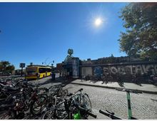 Ein städtischer Verkehrsknotenpunkt mit einem gelben Linienbus, Fahrrädern an Fahrradständern und Beschilderung für die S-Bahn und U-Bahn. Im Hintergrund stehen graffiti-bedeckte Wände, und die Szene ist bei klarem Himmel und Sonnenlicht aufgenommen.