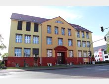 Gelbes und rotes Gebäude mit drei Stockwerken, großen rechteckigen Fenstern und einem roten Eingang mit der Beschriftung "Musik- und Kunstschule". Vor dem Gebäude befinden sich ein Gehweg, eine Rasenfläche und eine Straße, daneben ein Bereich mit kleinen Bäumen.