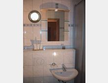 Kleines Badezimmer mit weißen Wandfliesen, einem Waschbecken, einem beleuchteten Spiegel mit Glasablagen und einer Duschkabine im rechten Bildbereich. Links befindet sich eine kleine, runde Wandlampe.