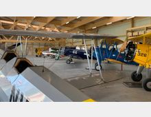 Ein Hangar mit mehreren historischen Doppeldecker-Flugzeugen, darunter ein gelbes, blaues und schwarzes Flugzeug. Die Holzkonstruktion des Dachs ist sichtbar, und der Boden des Hangars ist aus Beton.